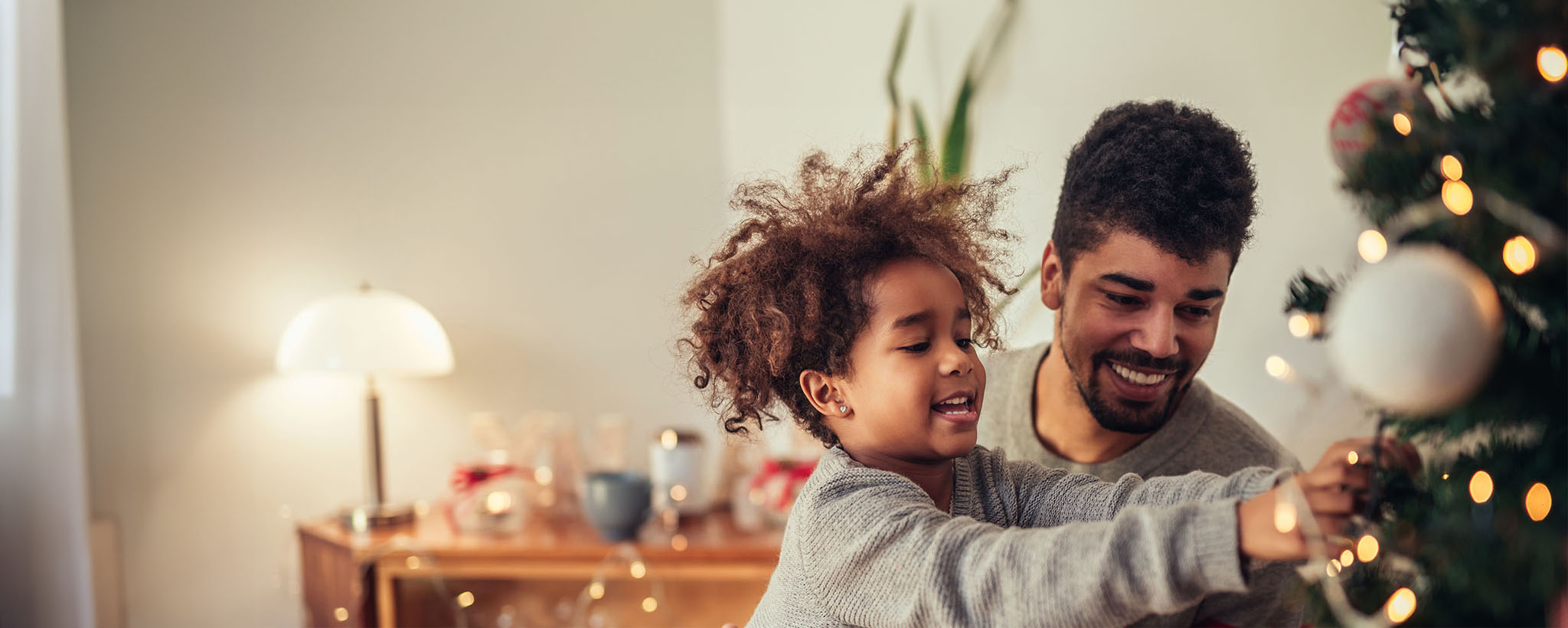 A father and school-aged daughter decorate a Christmas tree with lights and decorations. 