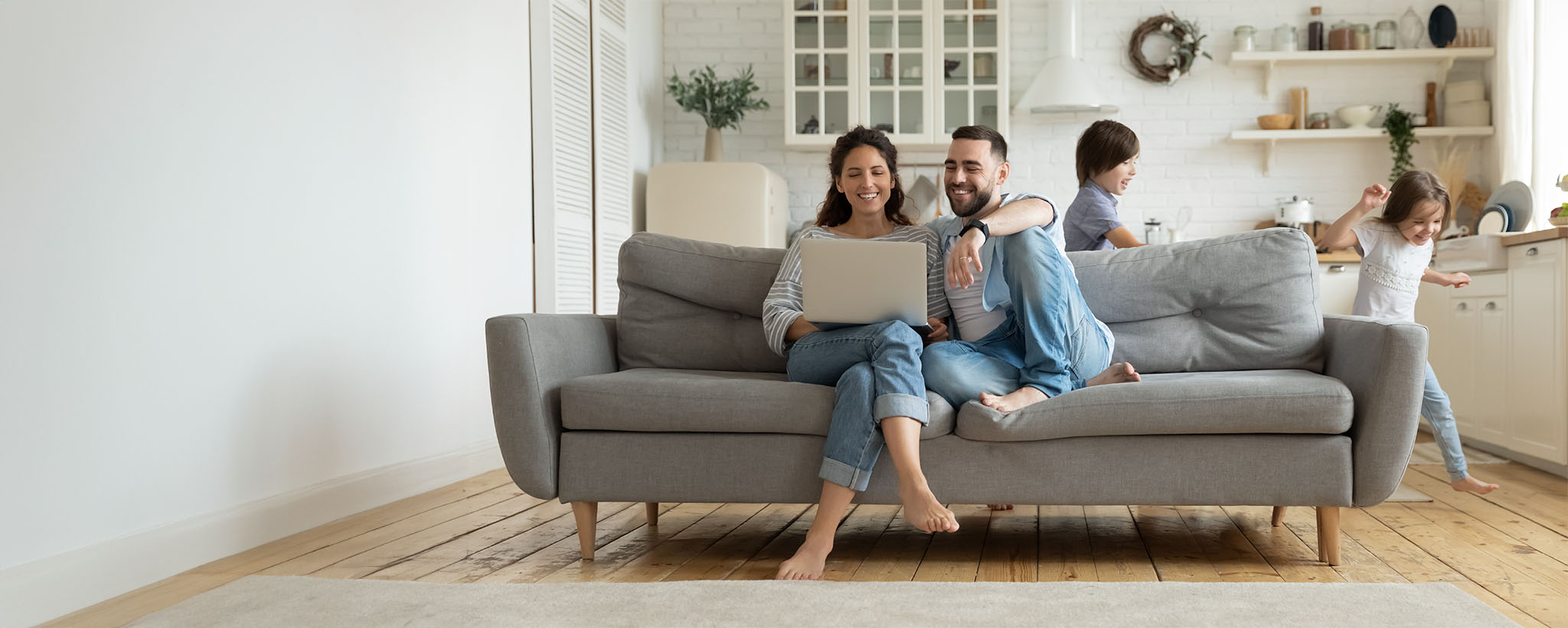 Una familia en su sala de estar. Una madre y un padre de mediana edad están sentados en el sofá mirando una computadora portátil mientras dos niños pequeños corren por ahí jugando.