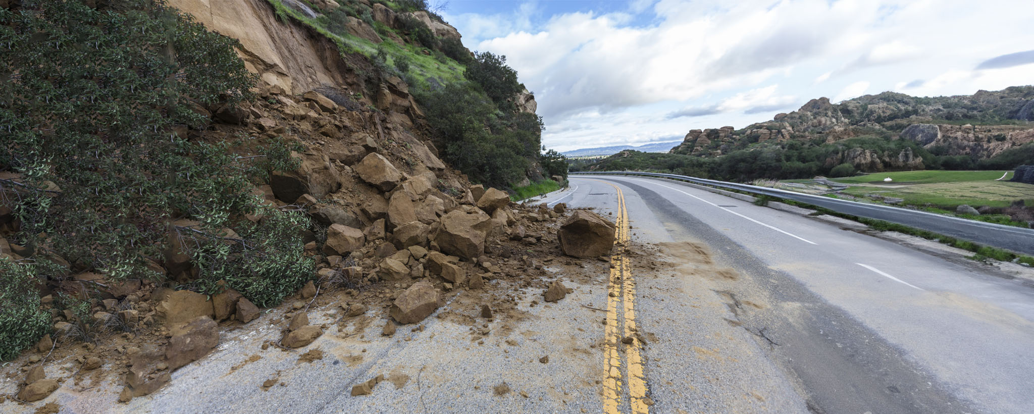 A landslide sends dirt and rocks tumbling onto the street, partially blocking the roadway.