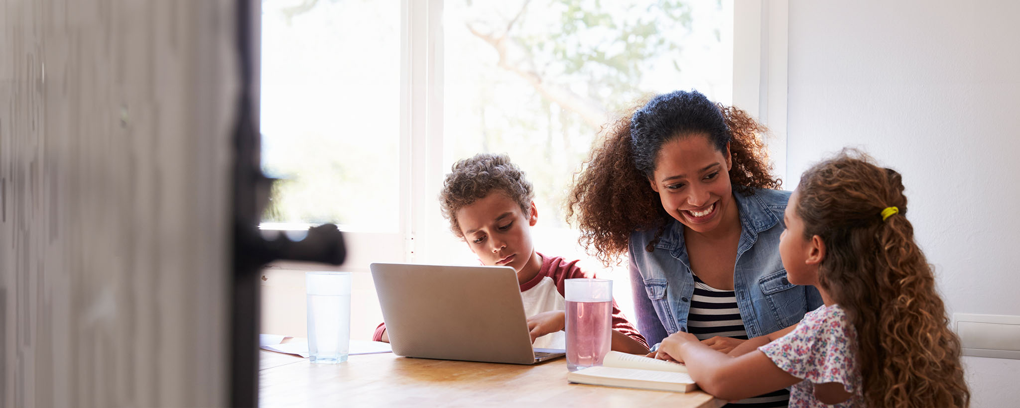 A mother sits with her school aged children at the kitchen table as they use a laptop and read a book. 