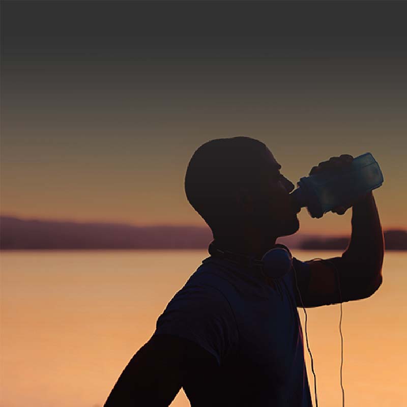 Photo of a man’s silhouette drinking from a water bottle near a lake as the sun sets. 