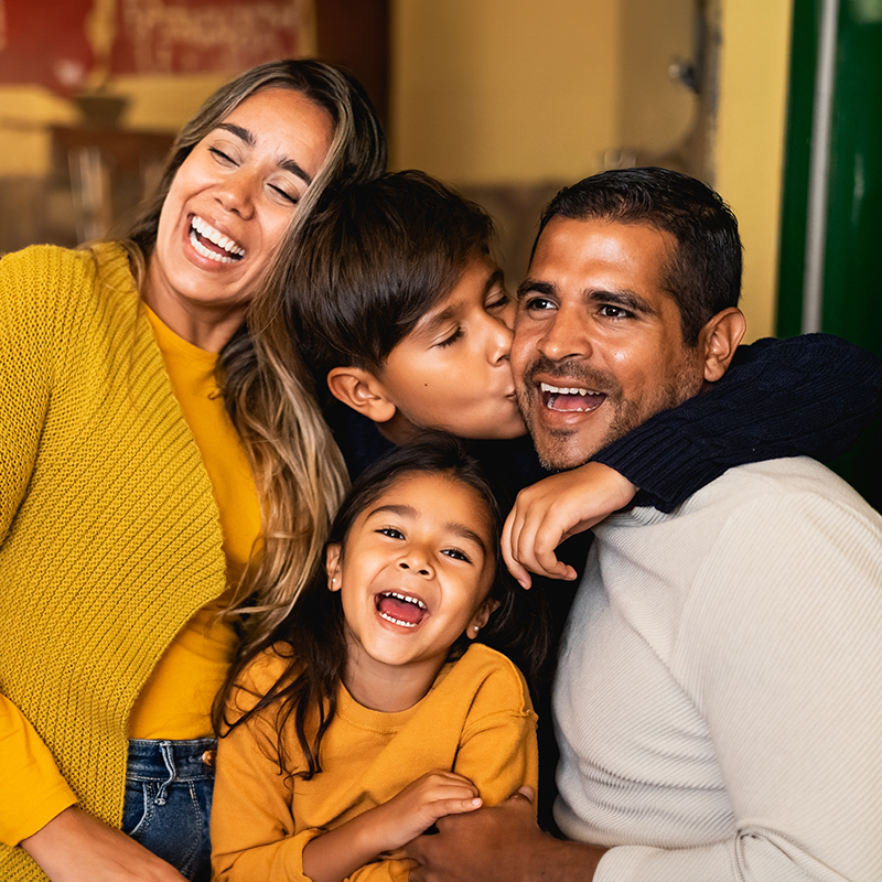 Una familia hispana compuesta por madre, padre, hijo e hija se abrazan juntos sonriendo.