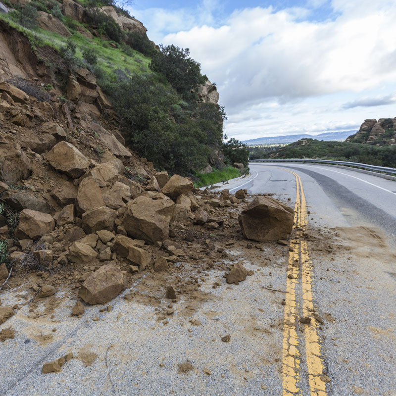 A landslide sends dirt and rocks tumbling onto the street, partially blocking the roadway.