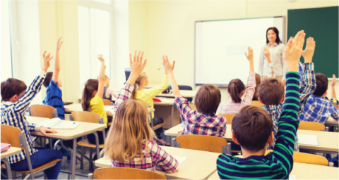 A teacher teaching a class. The children are raising their hands. 