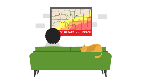 A woman sitting on the couch watching the news as it shows evacuation routes. A cat sits on the couch. 