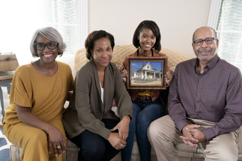 an african american family sits on a couch holding a picture of a home. 