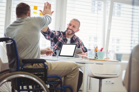 A man in a wheelchair at a table on his laptop, giving a high five to a friend. 